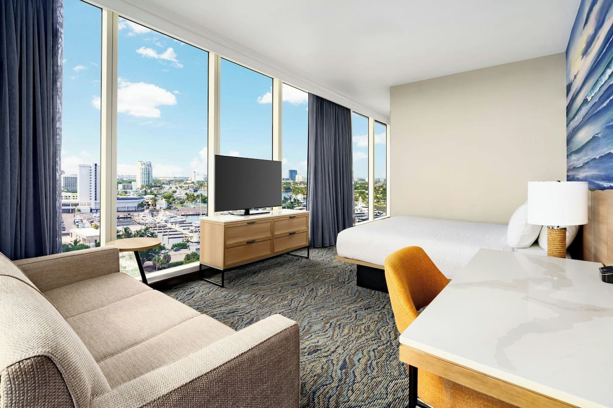 Ein Hotelzimmer mit großem Fenster, einem Bett, einer Couch, einem Schreibtisch und einem Fernseher.