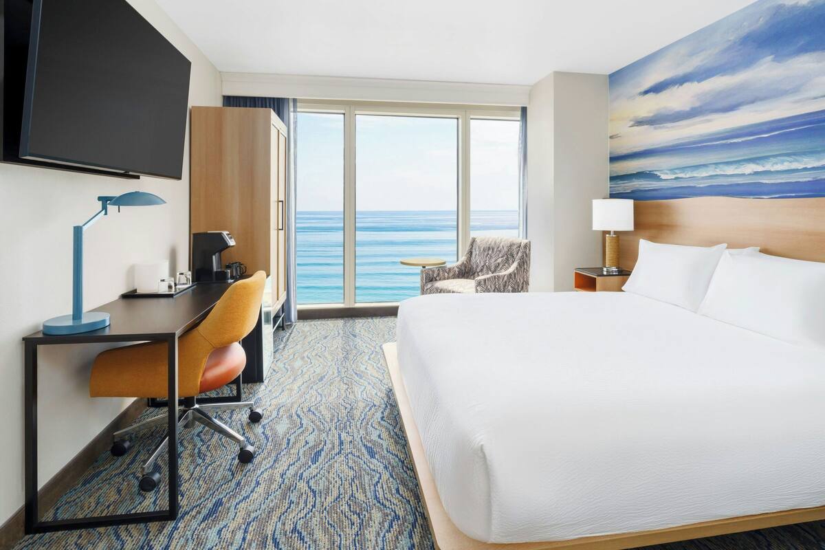 Ein Hotelzimmer mit einem großen Bett, einem Schreibtisch mit Stuhl, einem Fernseher und Blick auf den Ozean.