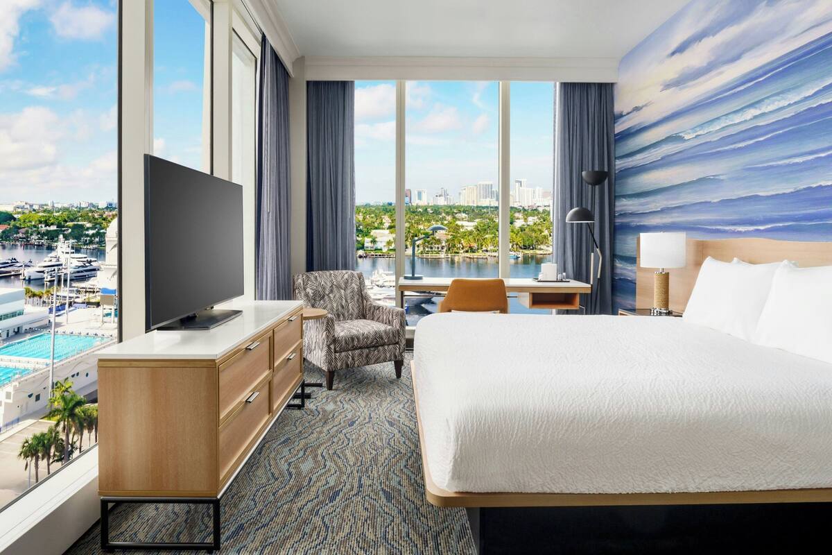 Ein Hotelzimmer mit einem großen Bett, einem Schreibtisch, einem Sessel, einem Fernseher und Blick auf einen Jachthafen und Stadtpanorama.