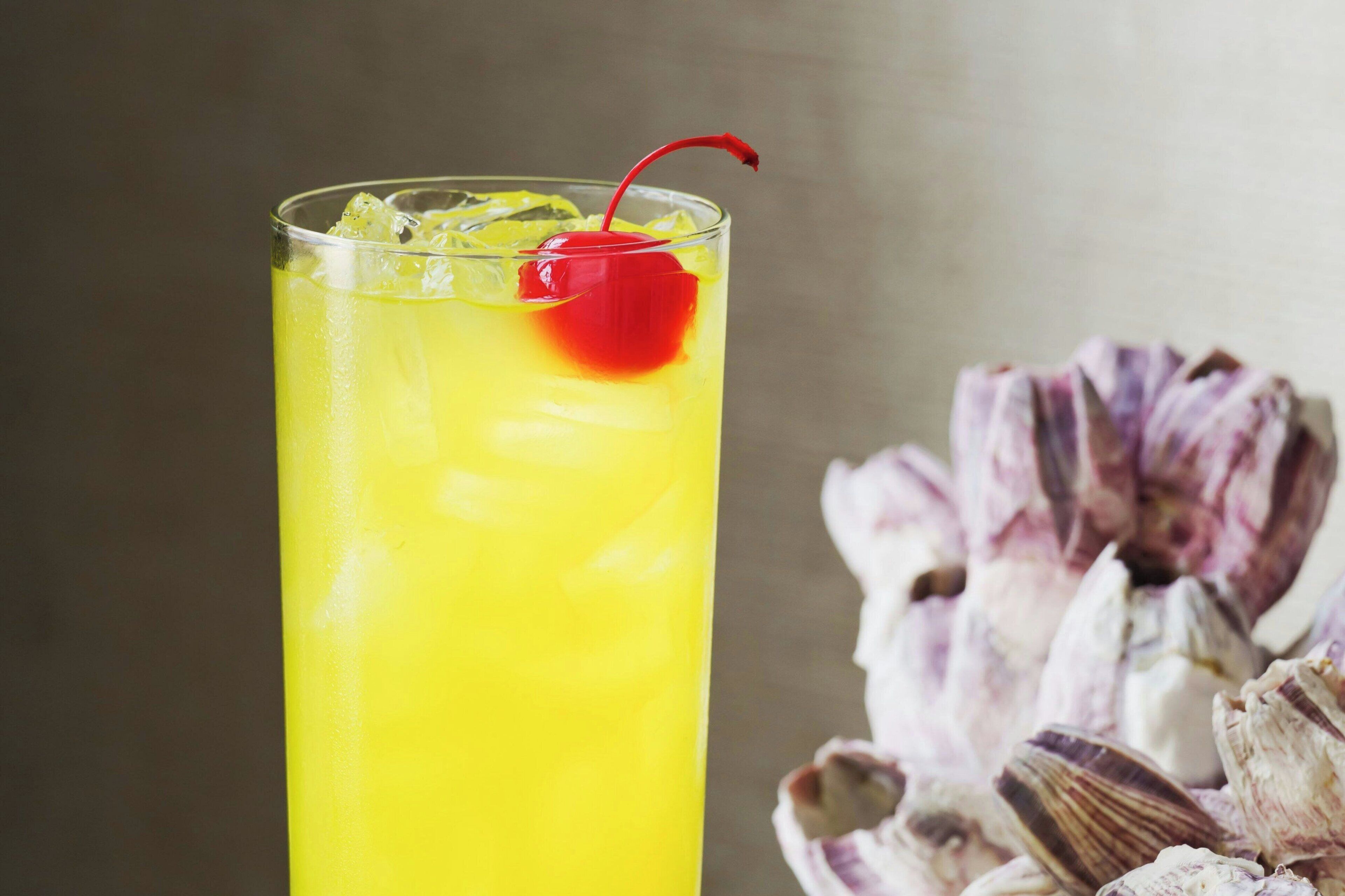 A tall glass with a yellow beverage, ice cubes, and a red cherry on top.