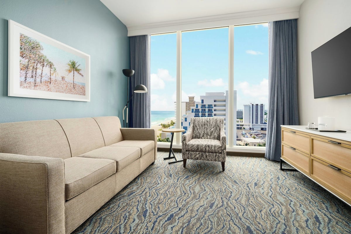 Ein Hotelzimmer mit großem Fenster, einer Couch, einem gemusterten Sessel, einem kleinen Tisch und einem Fernseher.