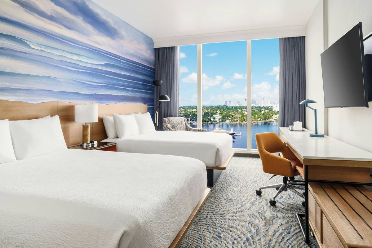 Ein Hotelzimmer mit zwei Betten, einem Schreibtisch, einem Stuhl, einem Fernseher und einem großen Fenster mit Blick auf die Stadt.
