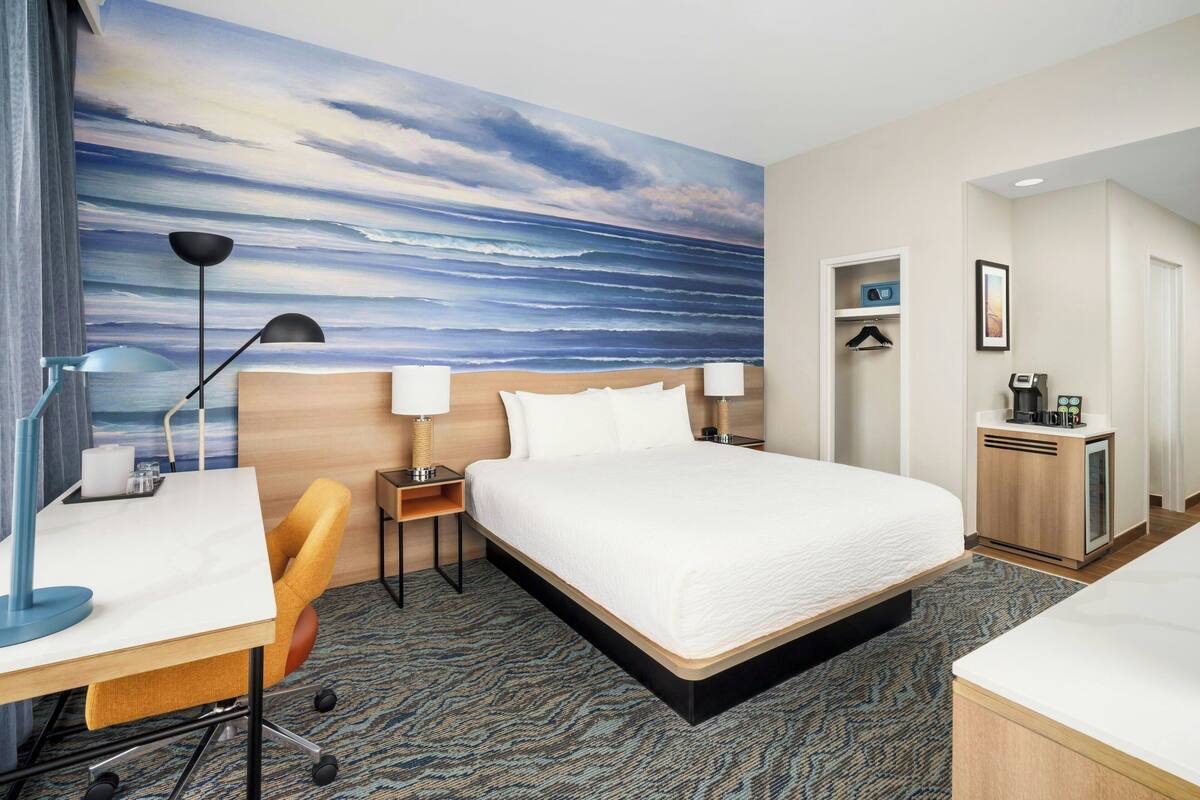 Ein Hotelzimmer mit einem großen Bett, einem Schreibtisch mit Stuhl, einer Schreibtischlampe, einem Nachttisch mit Lampe, einer Kaffeemaschine und einem Meereslandschaftsbild an der Wand.