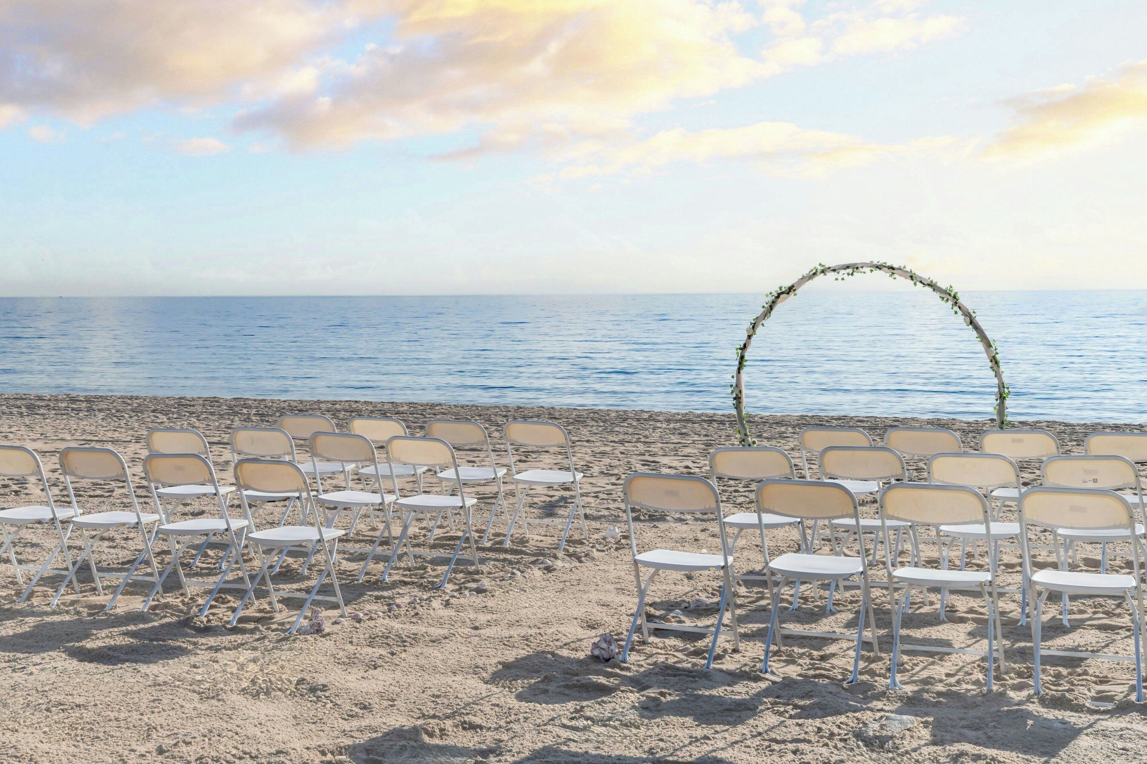 Ein Strandambiente mit Reihen aus weißen Klappstühlen, die für eine Veranstaltung aufgestellt sind, einem dekorativen Bogen aus Ästen und dem Meer im Hintergrund.