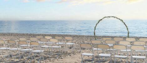 Ein Strandambiente mit Reihen aus weißen Klappstühlen, die für eine Veranstaltung aufgestellt sind, einem dekorativen Bogen aus Ästen und dem Meer im Hintergrund.