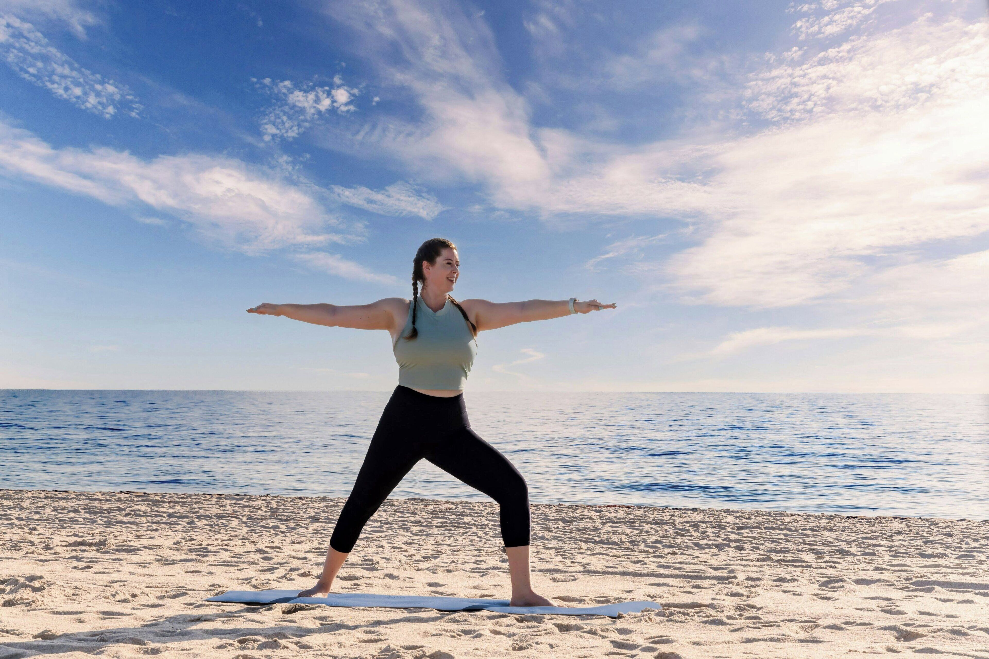 A person performing a yoga pose on a mat on a beach with the sea and sky in the background.
