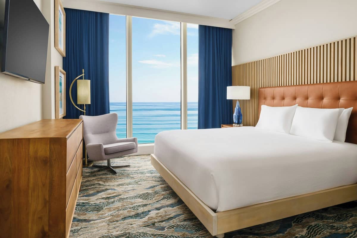 Ein Hotelzimmer mit einem großen Bett, einem Schreibtisch, einem Sessel und Blick aufs Meer.