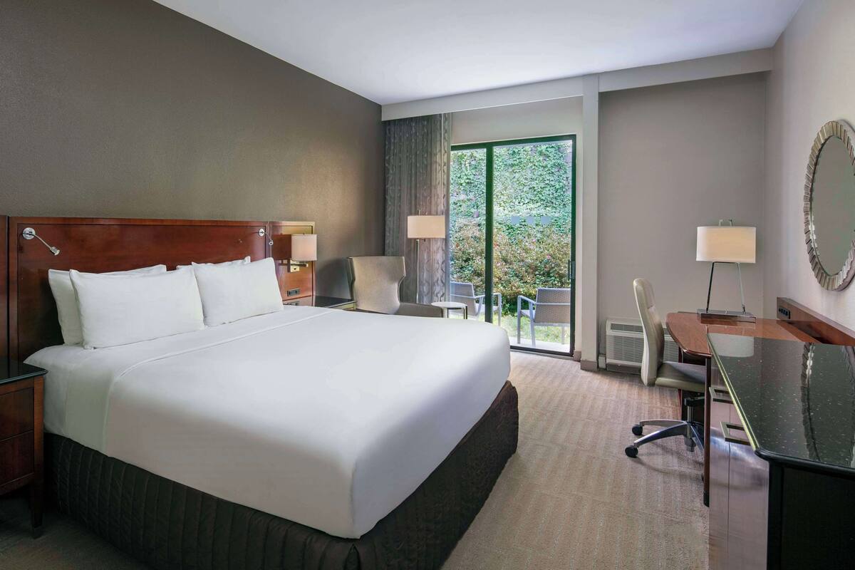 A hotel room with a large bed, a desk, a chair, a lamp, and a view of greenery through the window.
