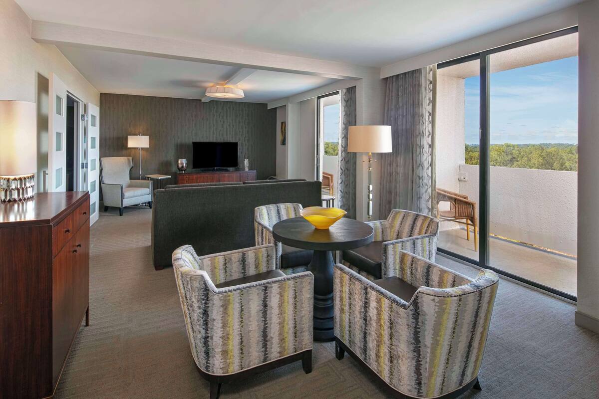 A modern hotel room with a balcony, a dining area with striped chairs, a flat-screen TV, and a wooden desk.