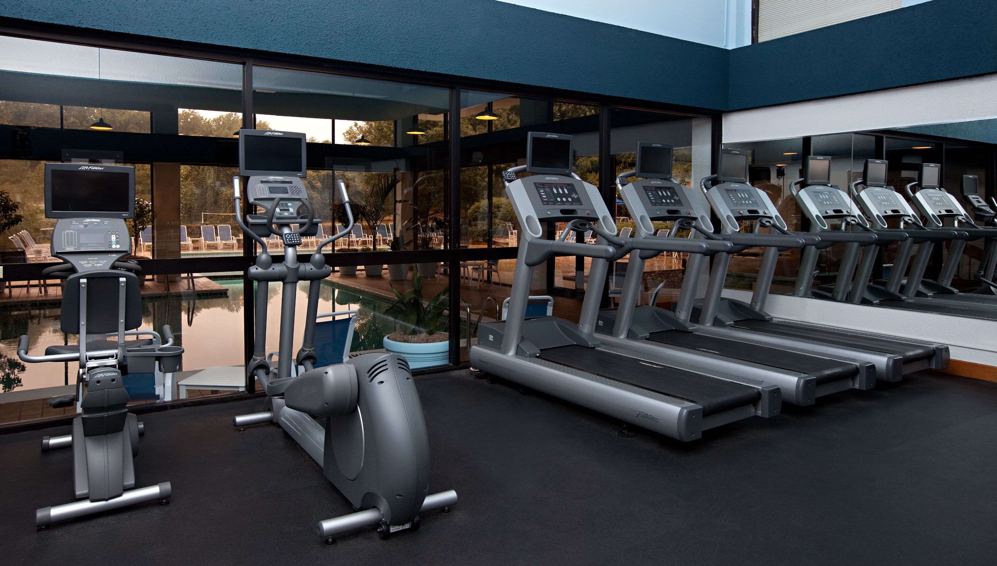 A modern gym with treadmills, an elliptical, and a rowing machine, featuring large windows and a pool view.