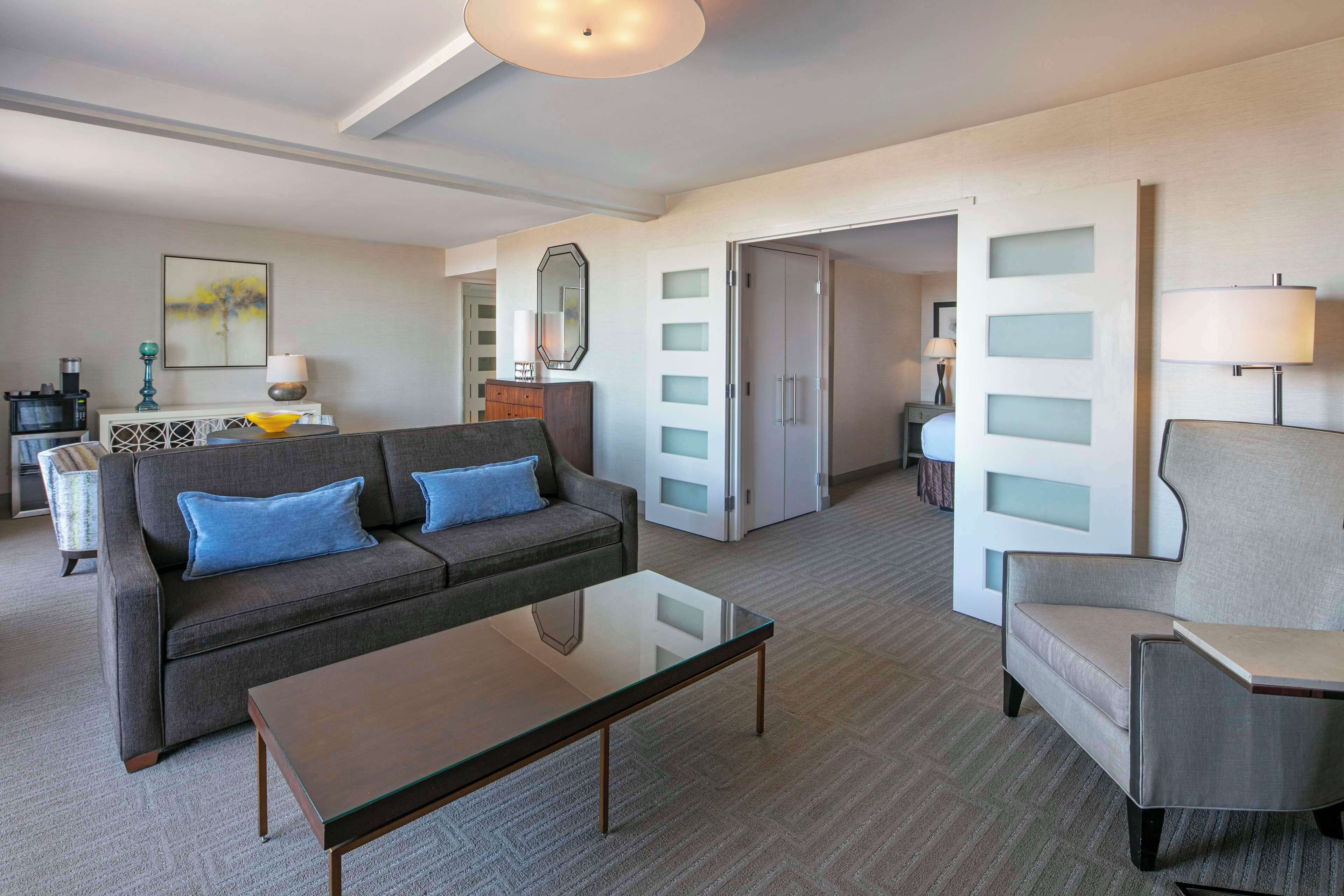 Ein modernes Hotelzimmer mit einem grauen Sofa, einem Glastisch und einem grauen Sessel. Es gibt einen Schreibtisch mit einer Lampe und einen Spiegel an der Wand.