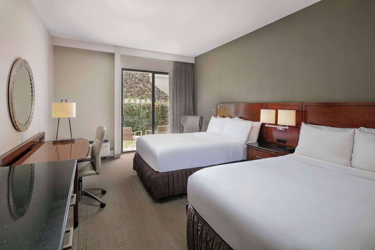 A hotel room with two beds, a desk, a chair, a lamp, and a view of the outdoors.