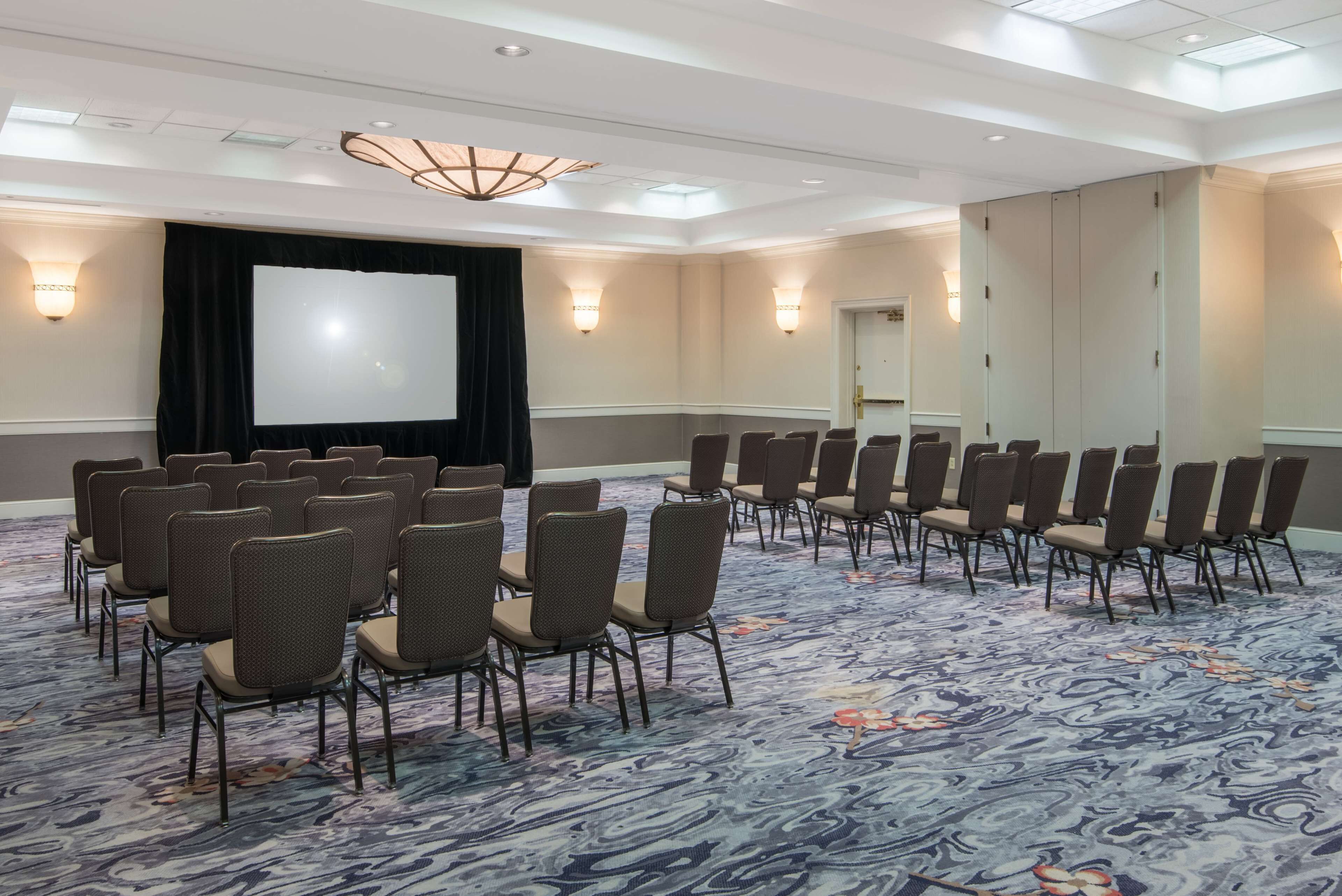 A conference room with rows of chairs, a large screen, and wall sconces.