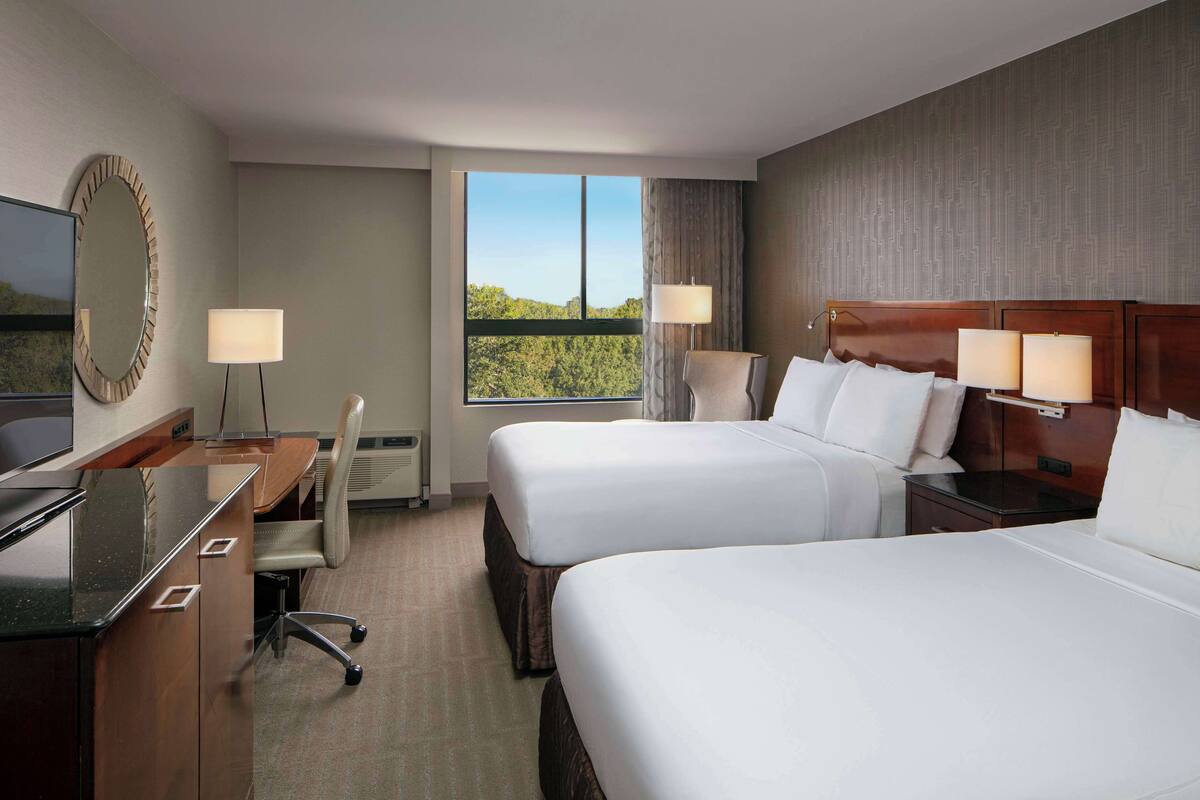 A hotel room with two beds, a desk, a chair, a TV, and a large window offering a view of the outdoors.