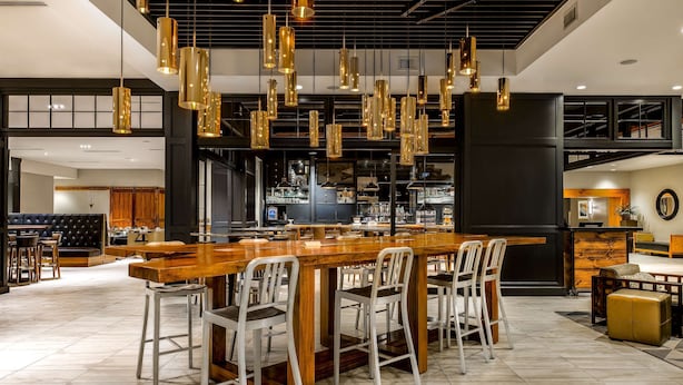 A spacious bar area with a long wooden counter and multiple bar stools, featuring pendant lighting and a modern industrial ceiling design. - Sonesta Charlotte Lower South End (Charlotte)