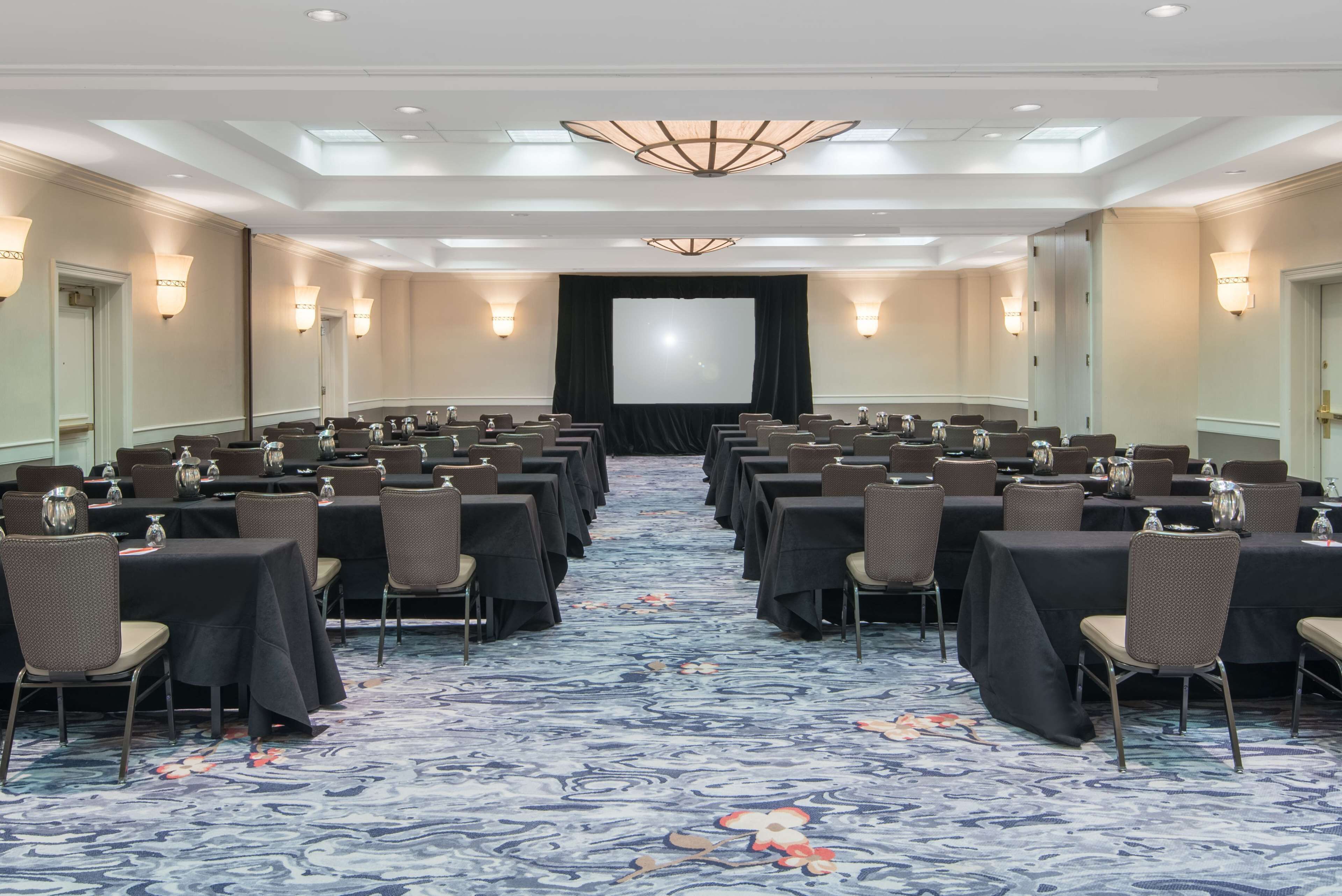 A conference room with rows of tables and chairs, a large screen at the front, and a patterned carpet.
