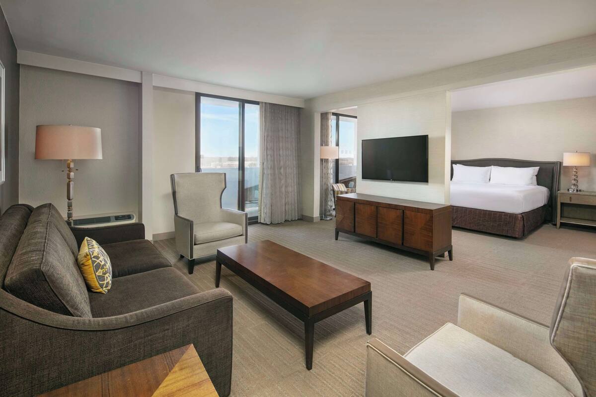 A hotel room with a sofa, armchair, bed, TV, and a view of the city.