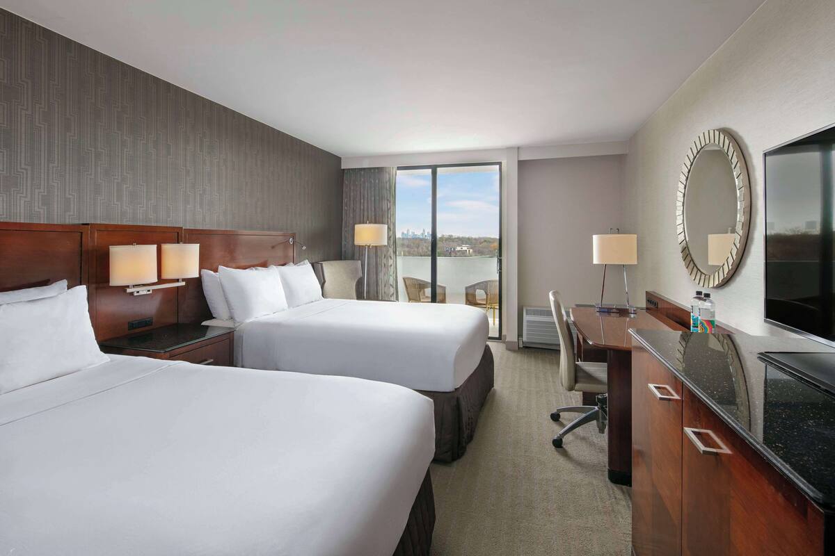 A hotel room with two beds, a desk, a chair, a TV, and a large window with a view.