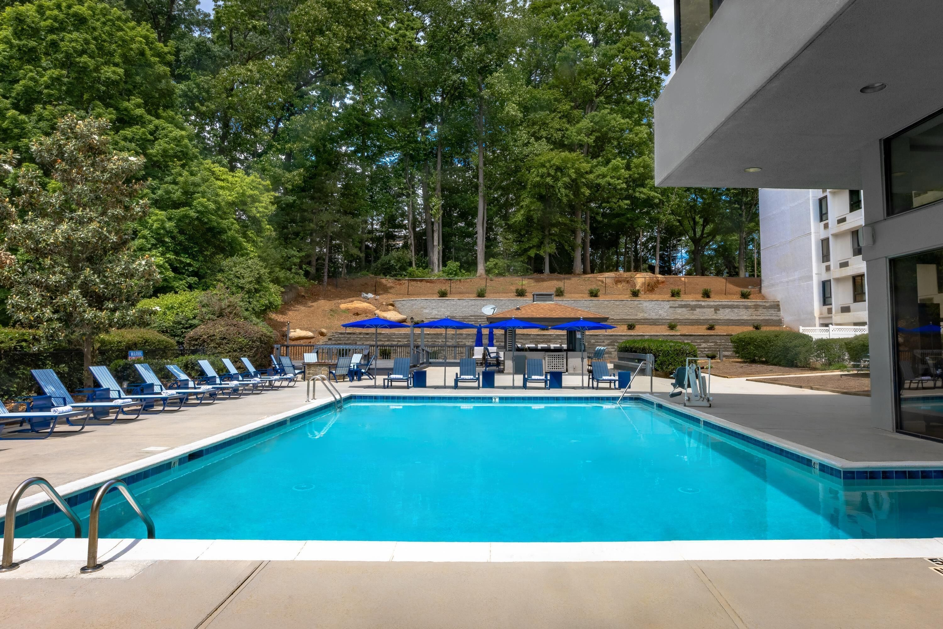 Una piscina con sillas y sombrillas azules, rodeada de árboles y un edificio.