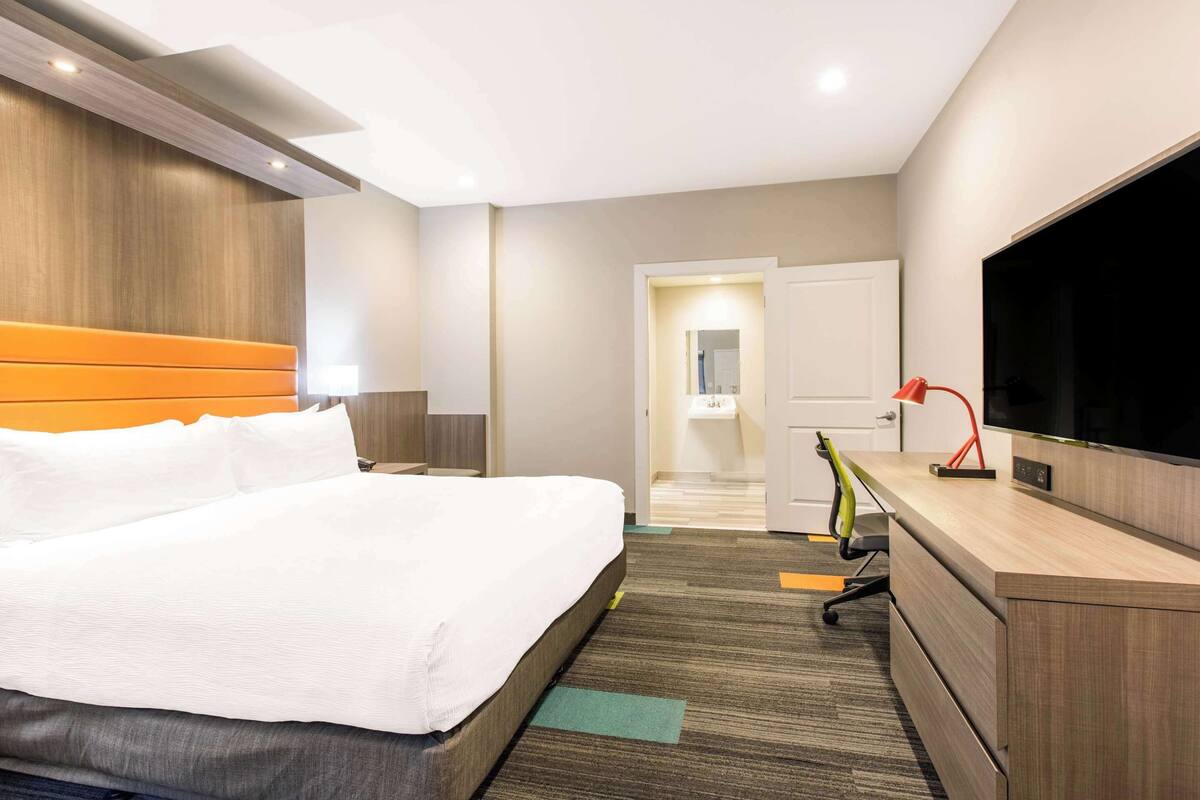 A hotel room with a bed, a desk, a TV, and a bathroom visible through an open door.