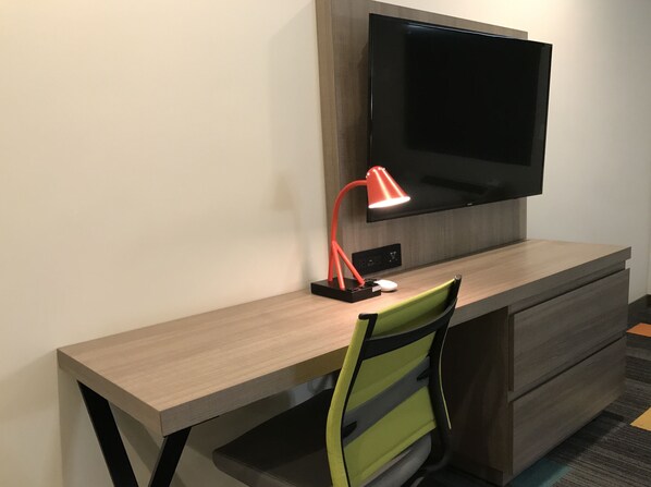 A modern hotel room with a wooden desk, a green chair, a flat-screen TV, and a red desk lamp.
