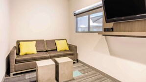 A modern living room with a grey sofa, two yellow cushions, a flat-screen TV, and a window with blinds. - Rodeway Inn near Melrose Ave (Los Angeles)