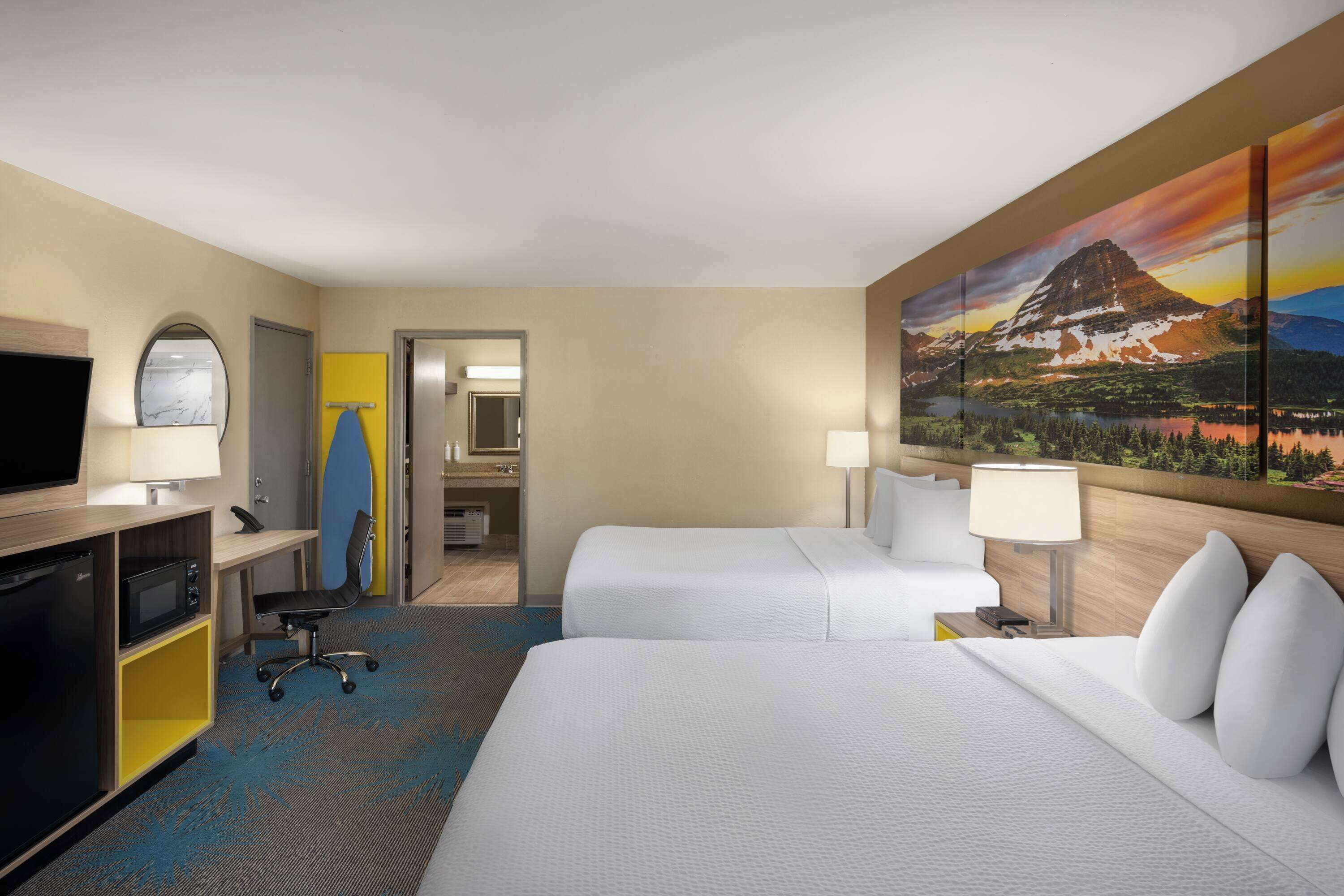 A hotel room with two beds, a desk with a computer, a TV, and a painting of a mountain landscape.