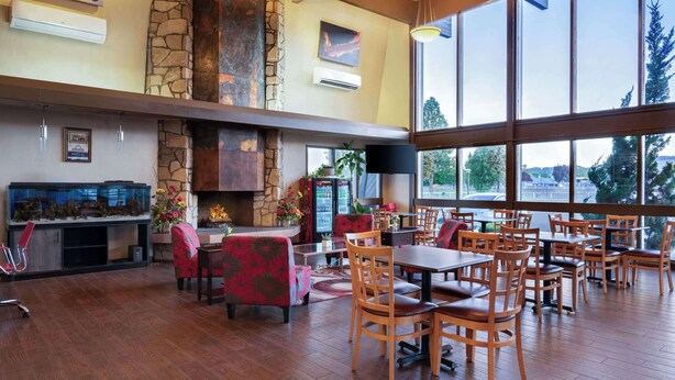 A spacious indoor area with wooden flooring, multiple tables and chairs, a stone fireplace, and large windows offering a view of the outdoors.