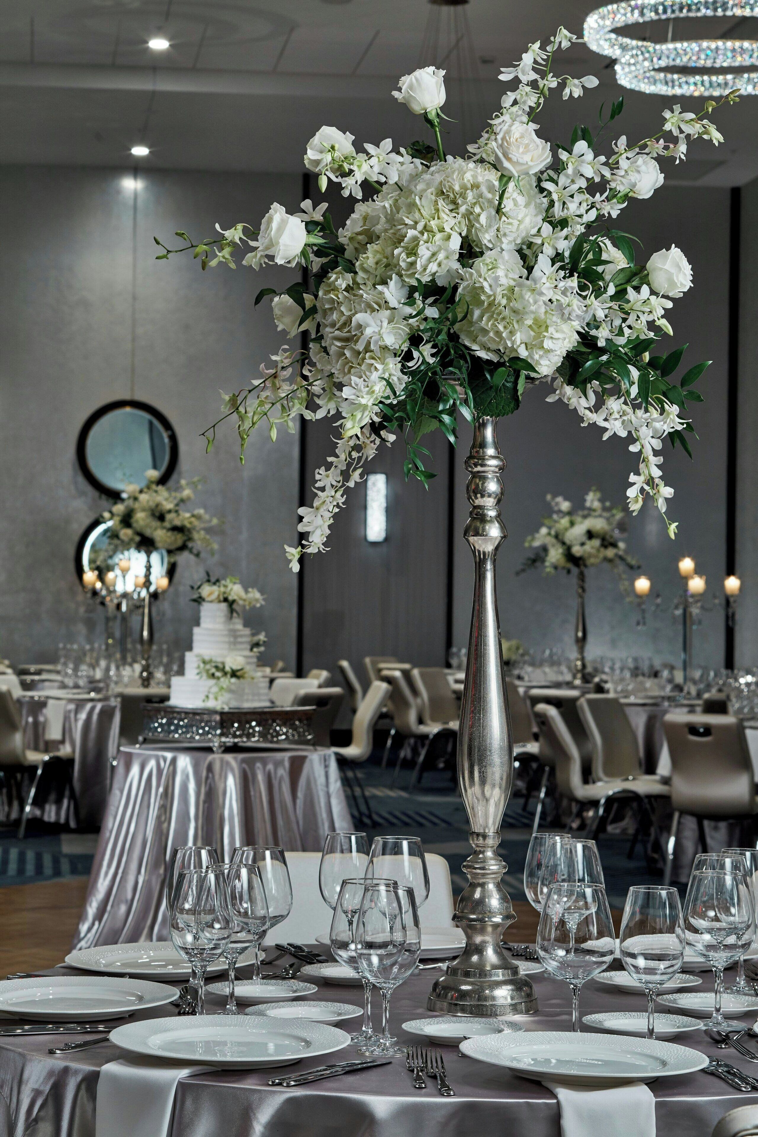 Een balzaal met gedekte tafels voor een formeel evenement, met hoge vazen met witte bloemen en een kroonluchter op de achtergrond.