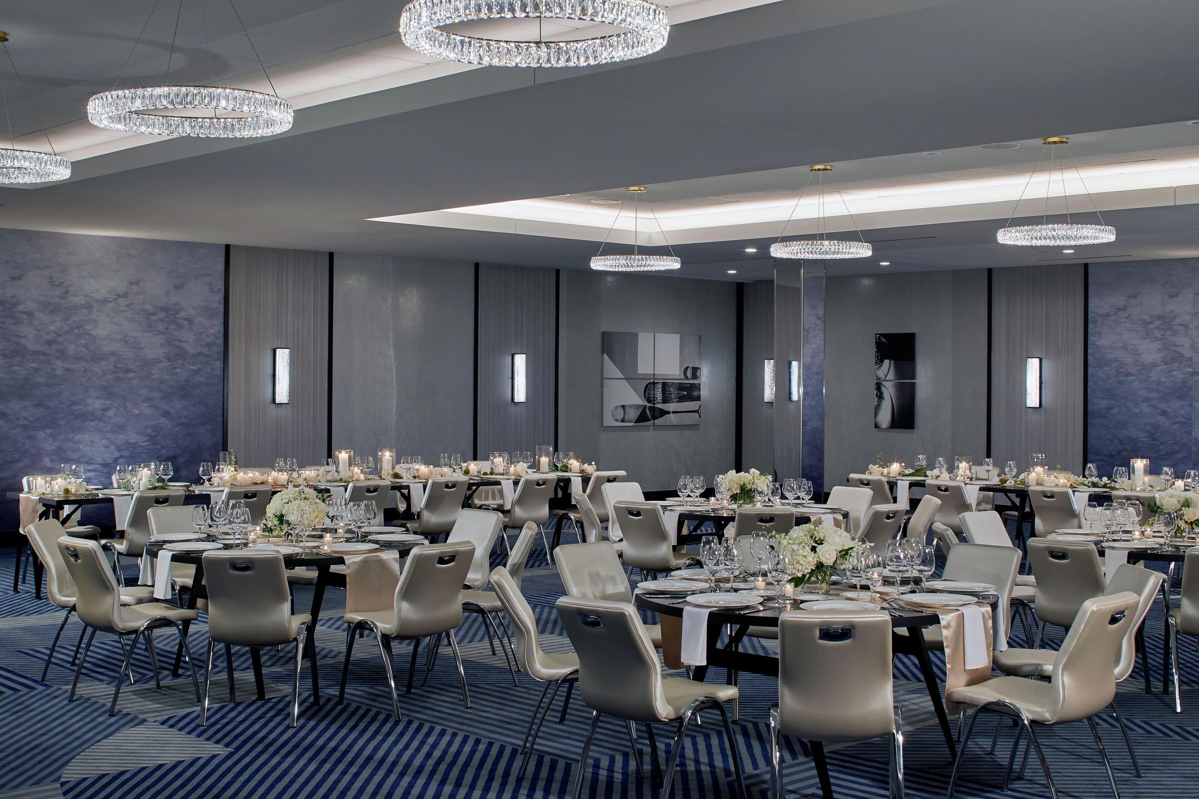 A large, well-lit banquet hall with multiple tables set for dining, featuring modern chairs and elegant chandeliers.