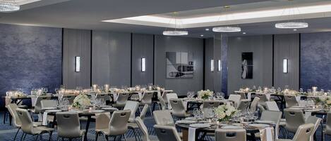 A large, well-lit banquet hall with multiple tables set for dining, featuring modern chairs and elegant chandeliers.
