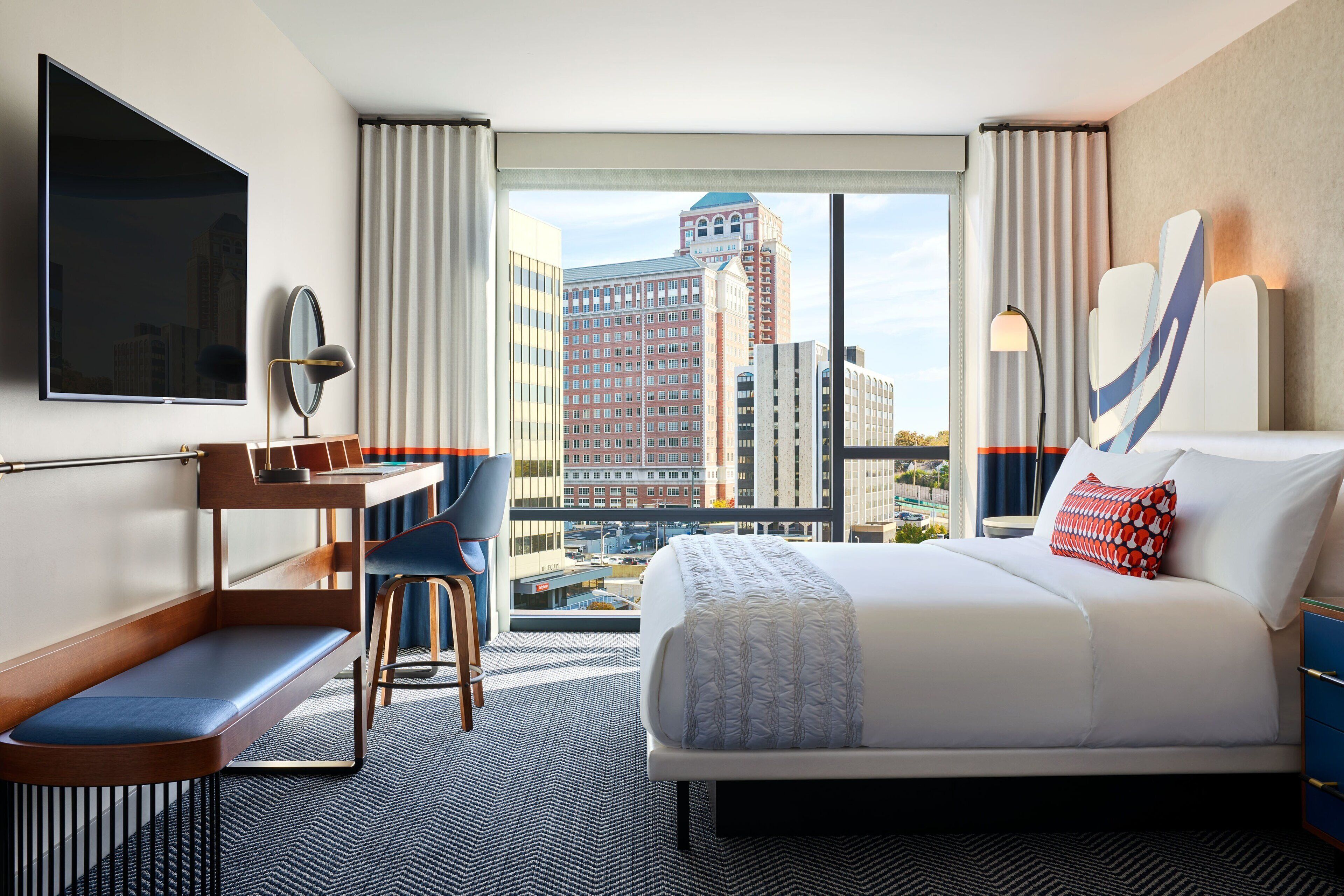 Een hotelkamer met een bed, bureau, stoel en een groot raam met uitzicht op de stad.
