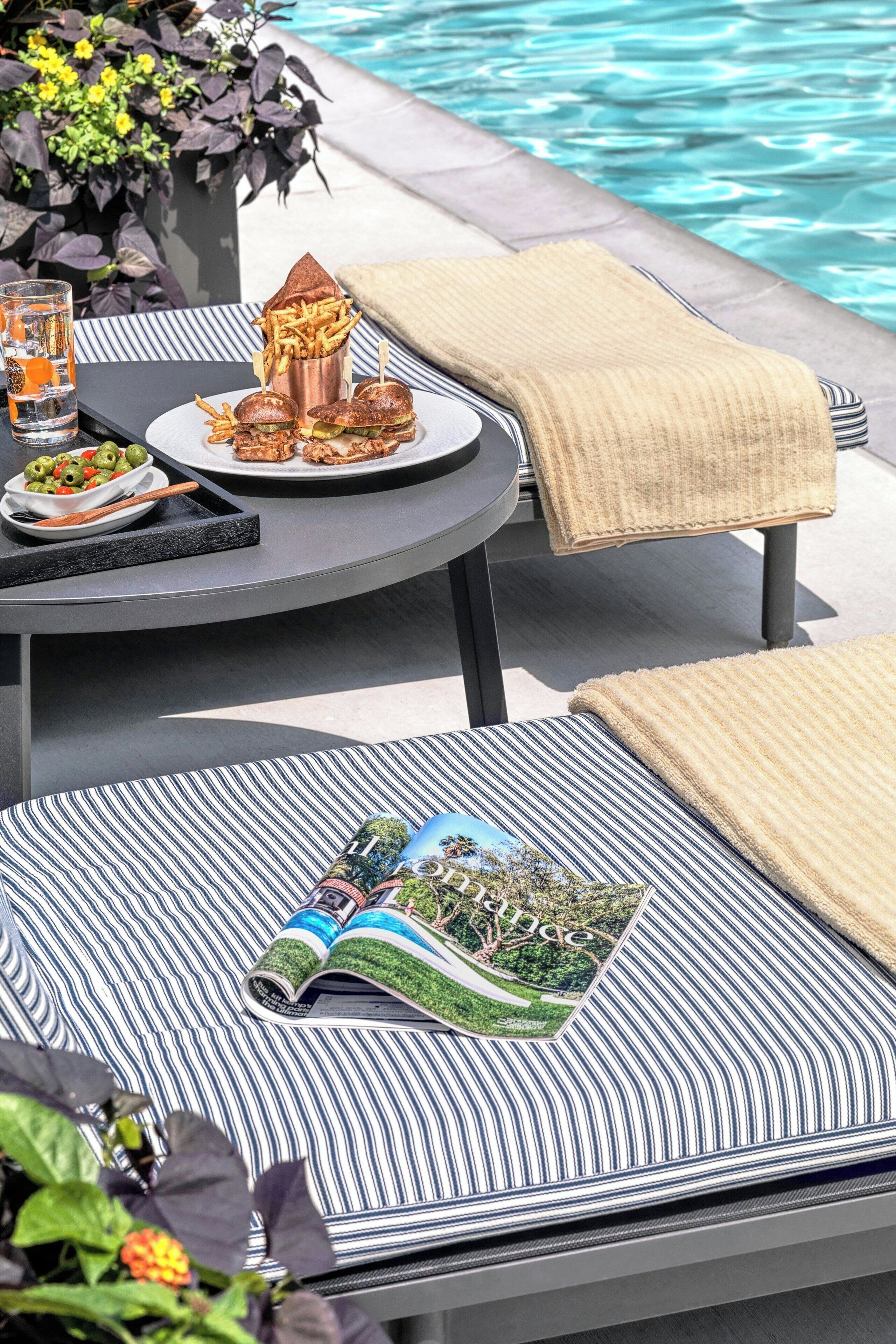 A poolside dining area with a table set for two, featuring a plate of food, a drink, and a magazine.