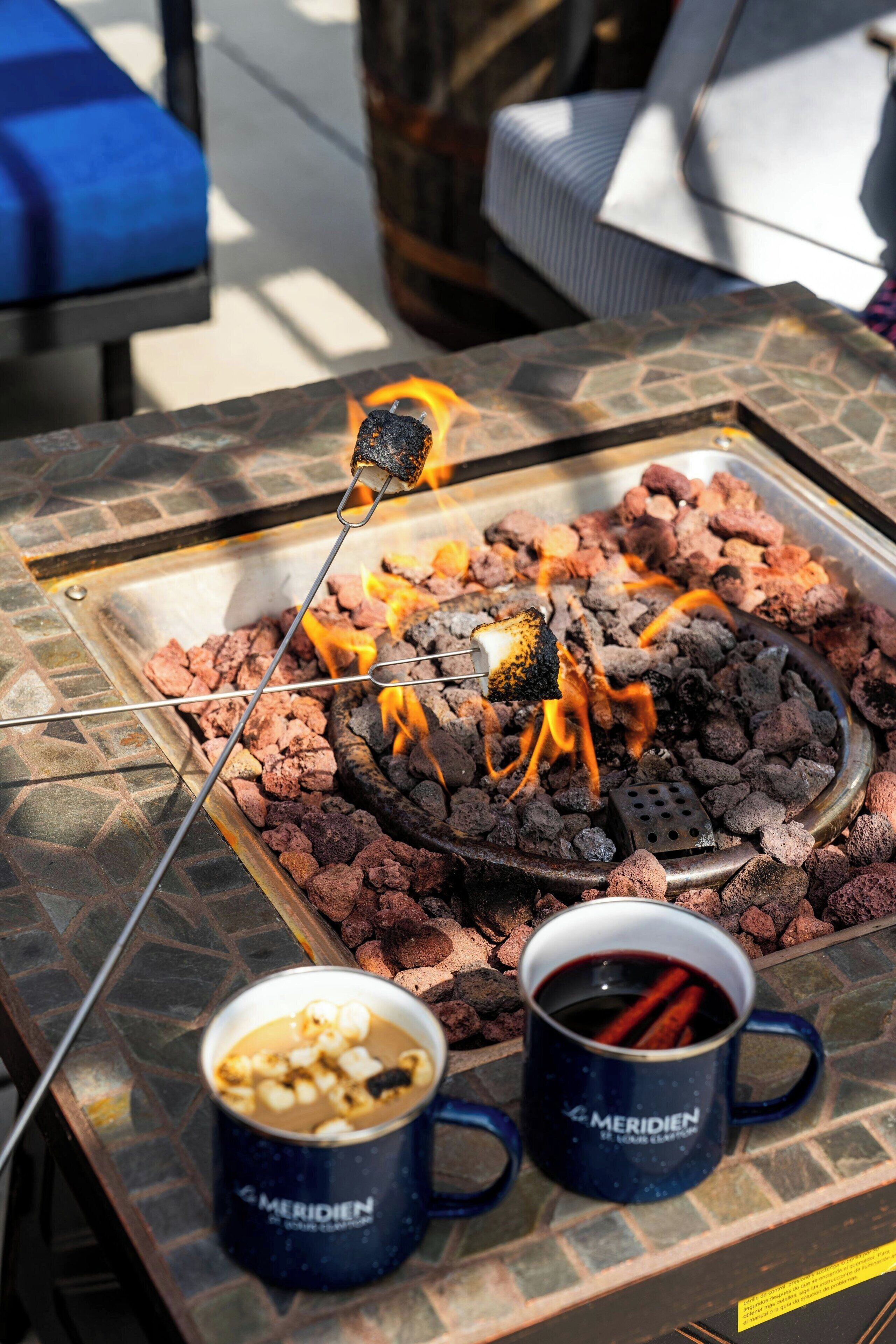 A charcoal grill with a skewer of food, two mugs, and a stone-tiled table.