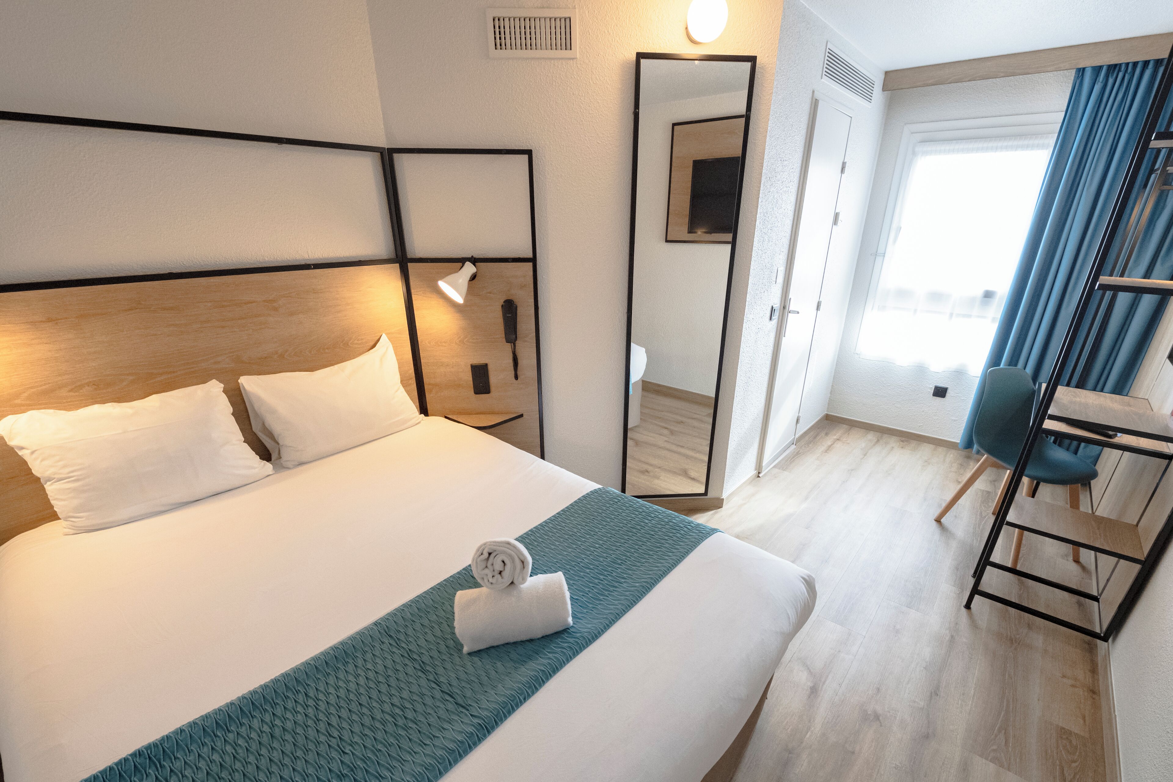 Une chambre d’hôtel avec un grand lit, un bureau, une chaise, un miroir et une fenêtre avec des rideaux.