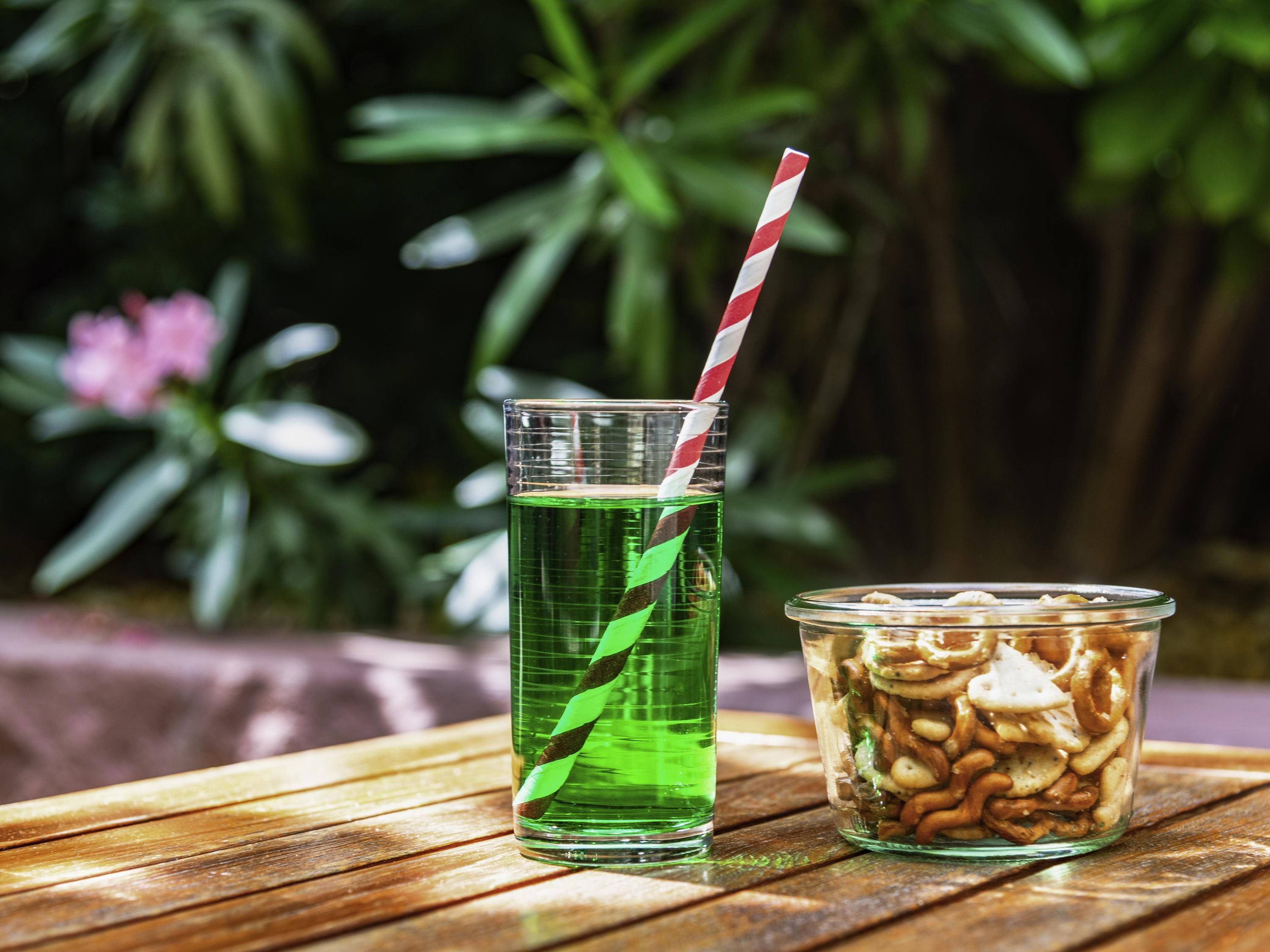 Un vaso de bebida verde con una pajita a rayas y un recipiente con frutos secos mezclados sobre una mesa de madera.