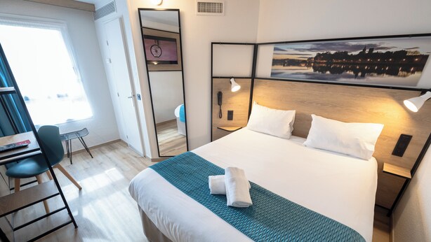 A hotel room with a bed, bedside table, desk, chair, and a large window. - ibis Avignon Centre Pont de l'Europe (Avignon)
