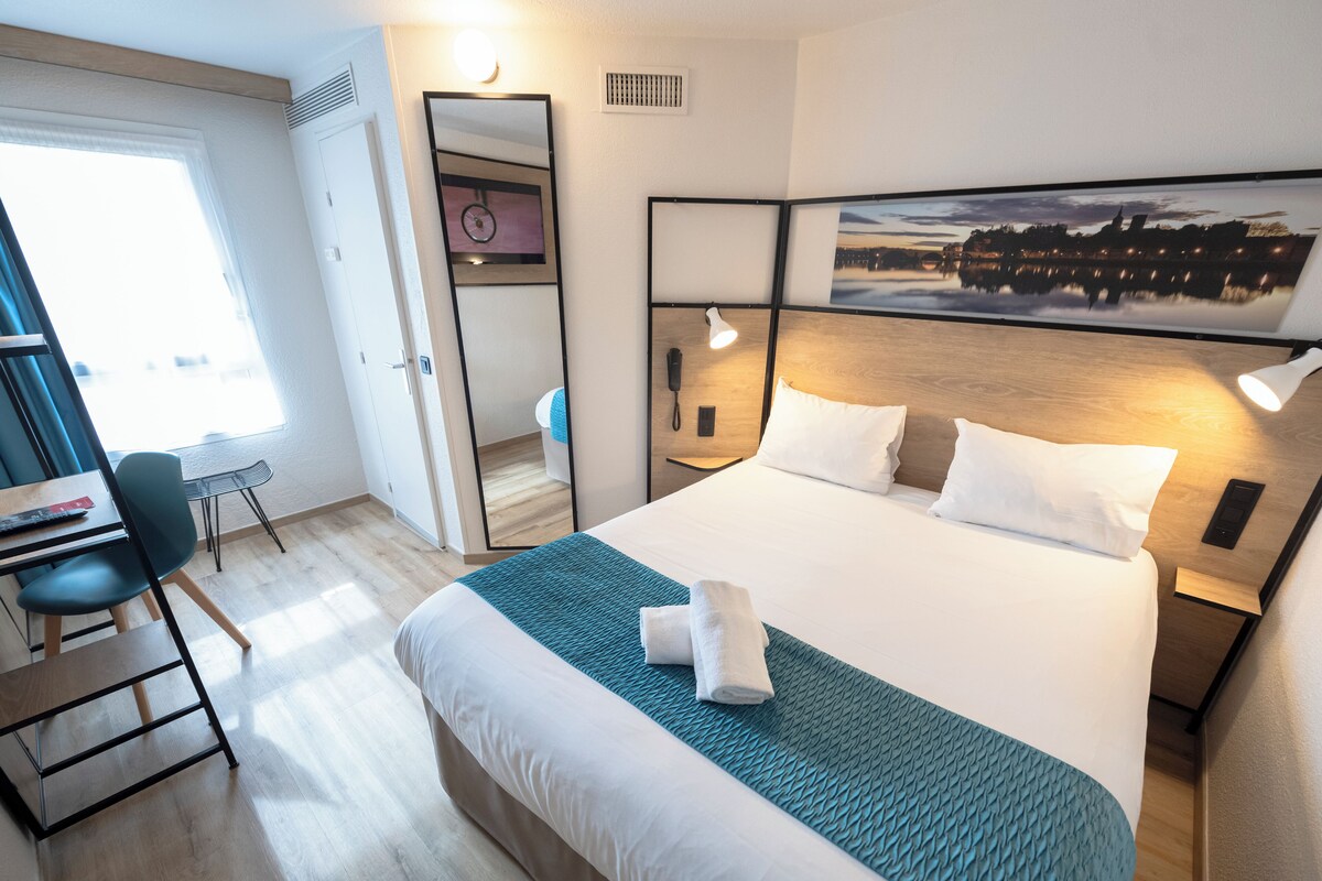 A hotel room with a bed, bedside table, desk, chair, and a large window.