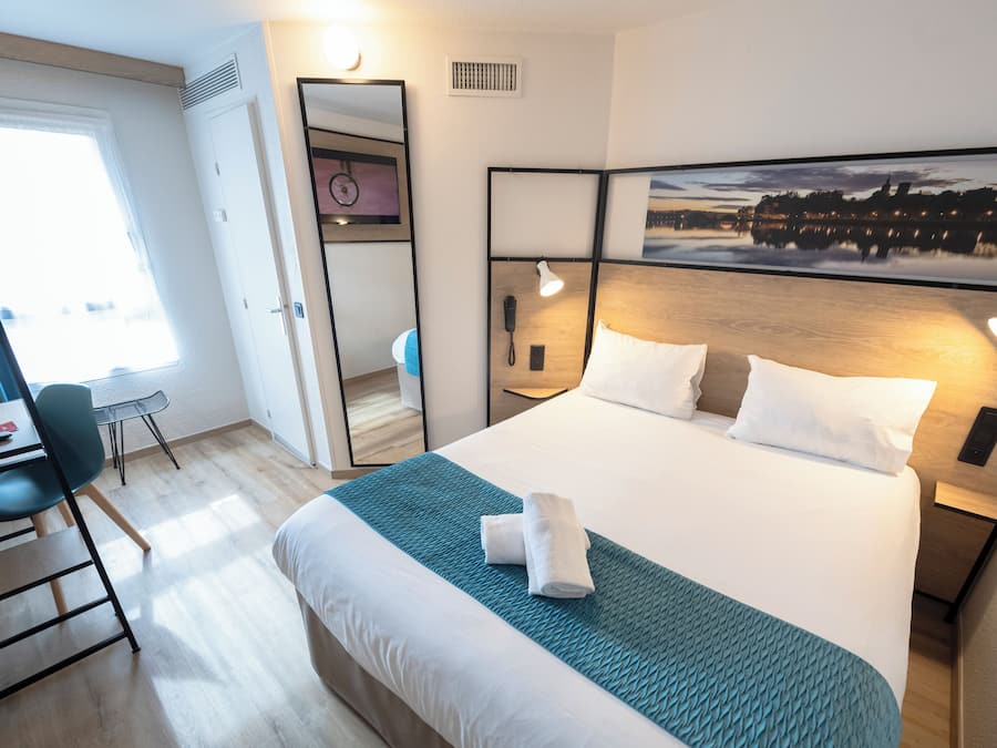 Une chambre d’hôtel avec un lit, une table de chevet, un bureau, une chaise et une grande fenêtre.