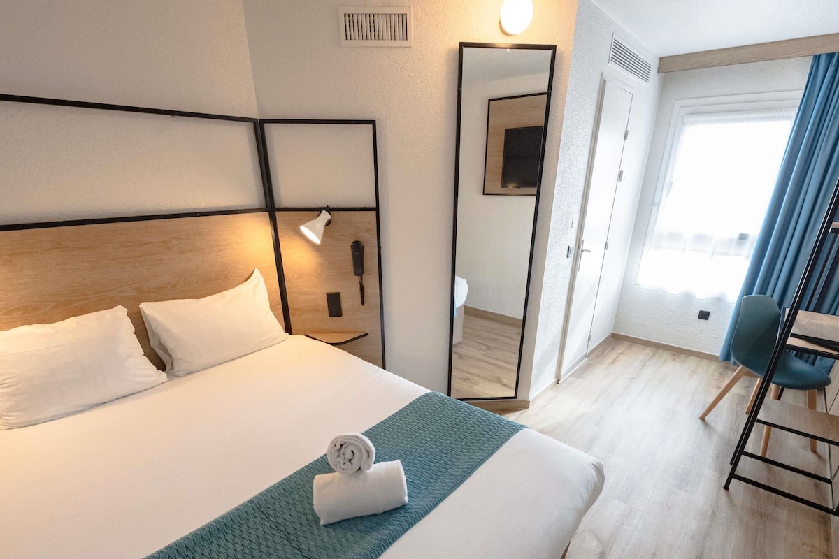 A hotel room with a bed, a desk, a chair, a mirror, and a window with curtains.