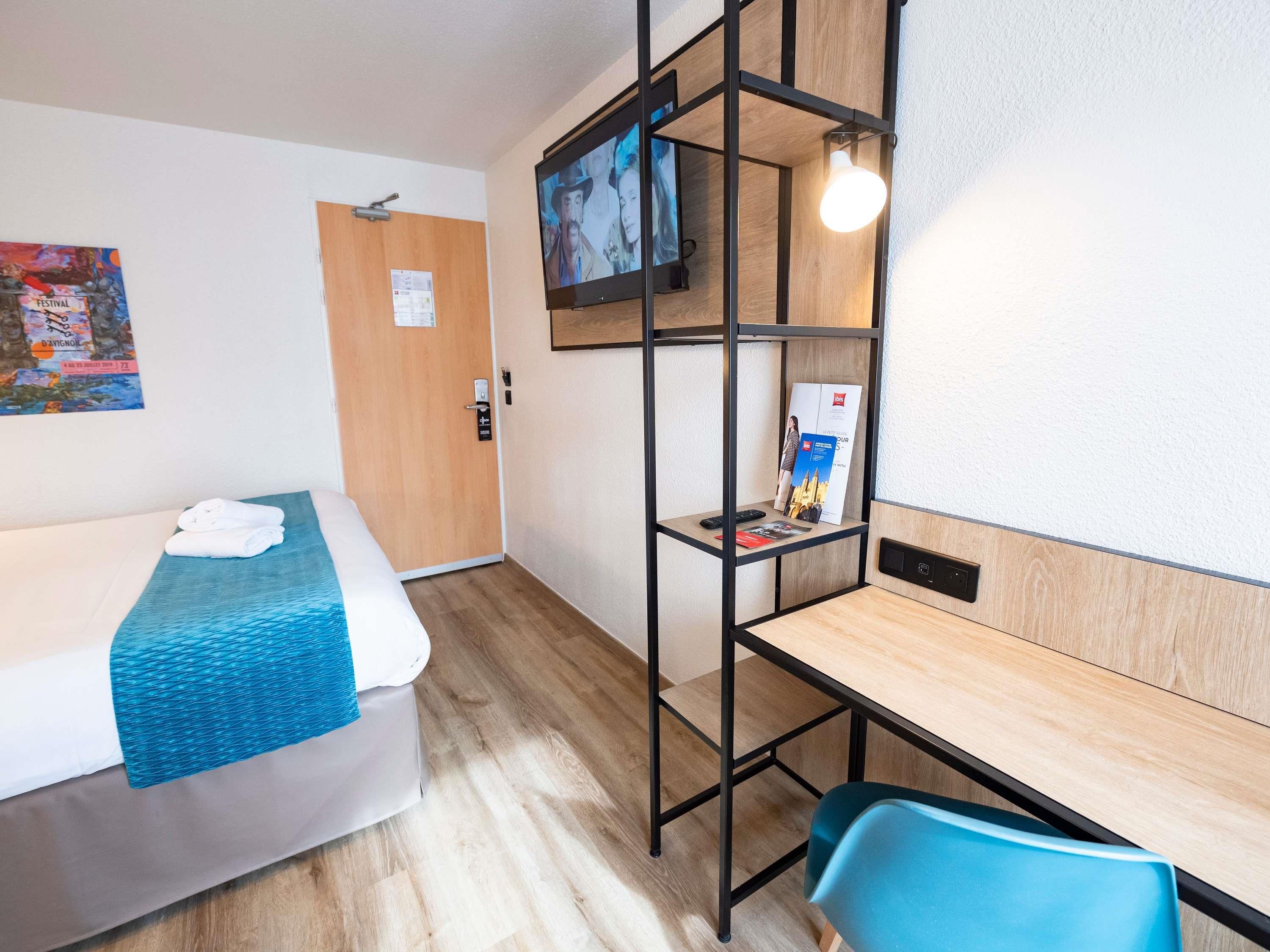 A hotel room with a bed, a desk, a chair, a TV, and a shelf with a magazine.