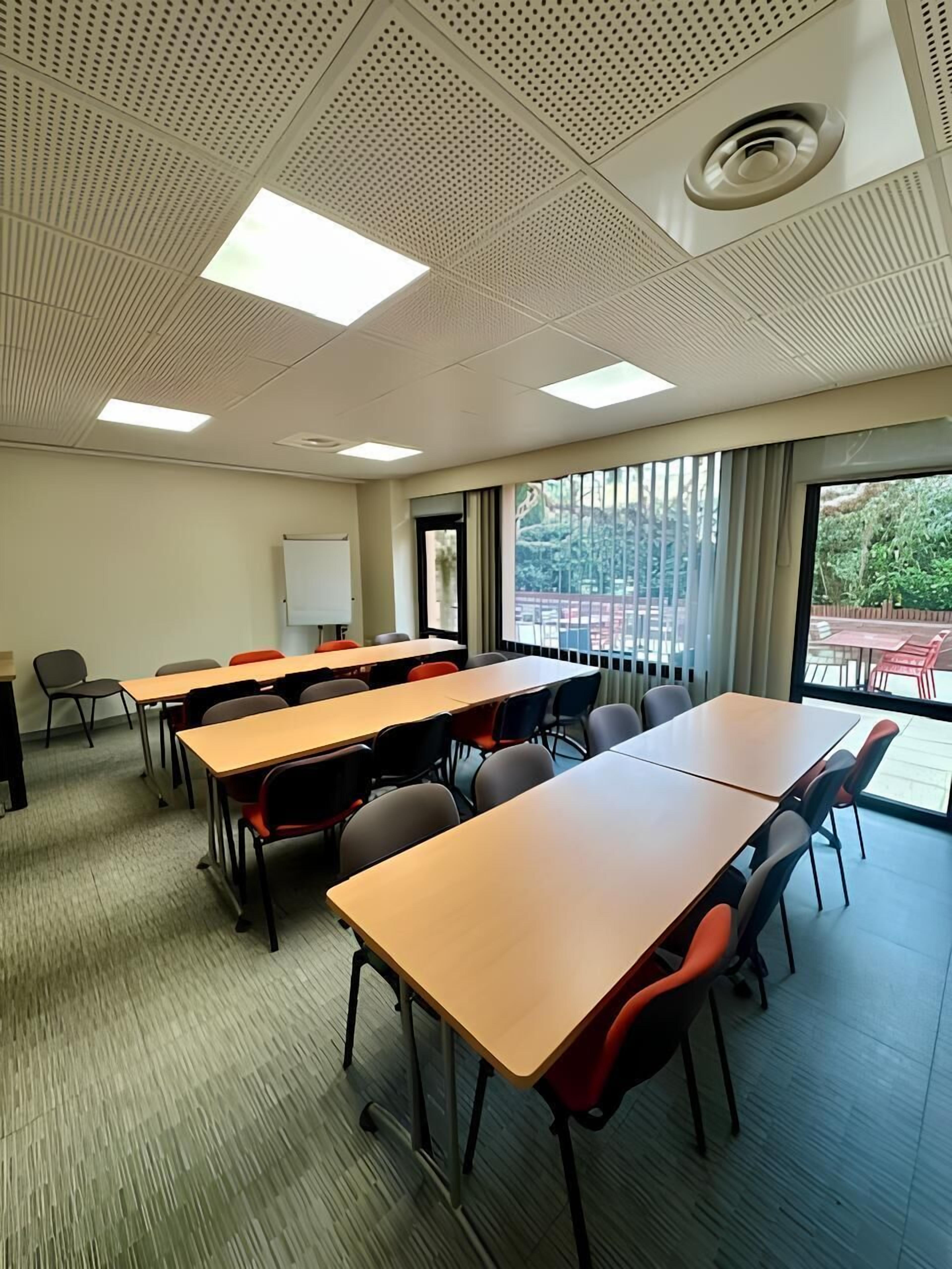 A conference room with multiple tables and chairs, a whiteboard, and large windows with a view of the outdoors.