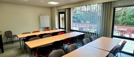A conference room with multiple tables and chairs, a whiteboard, and large windows with a view of the outdoors.