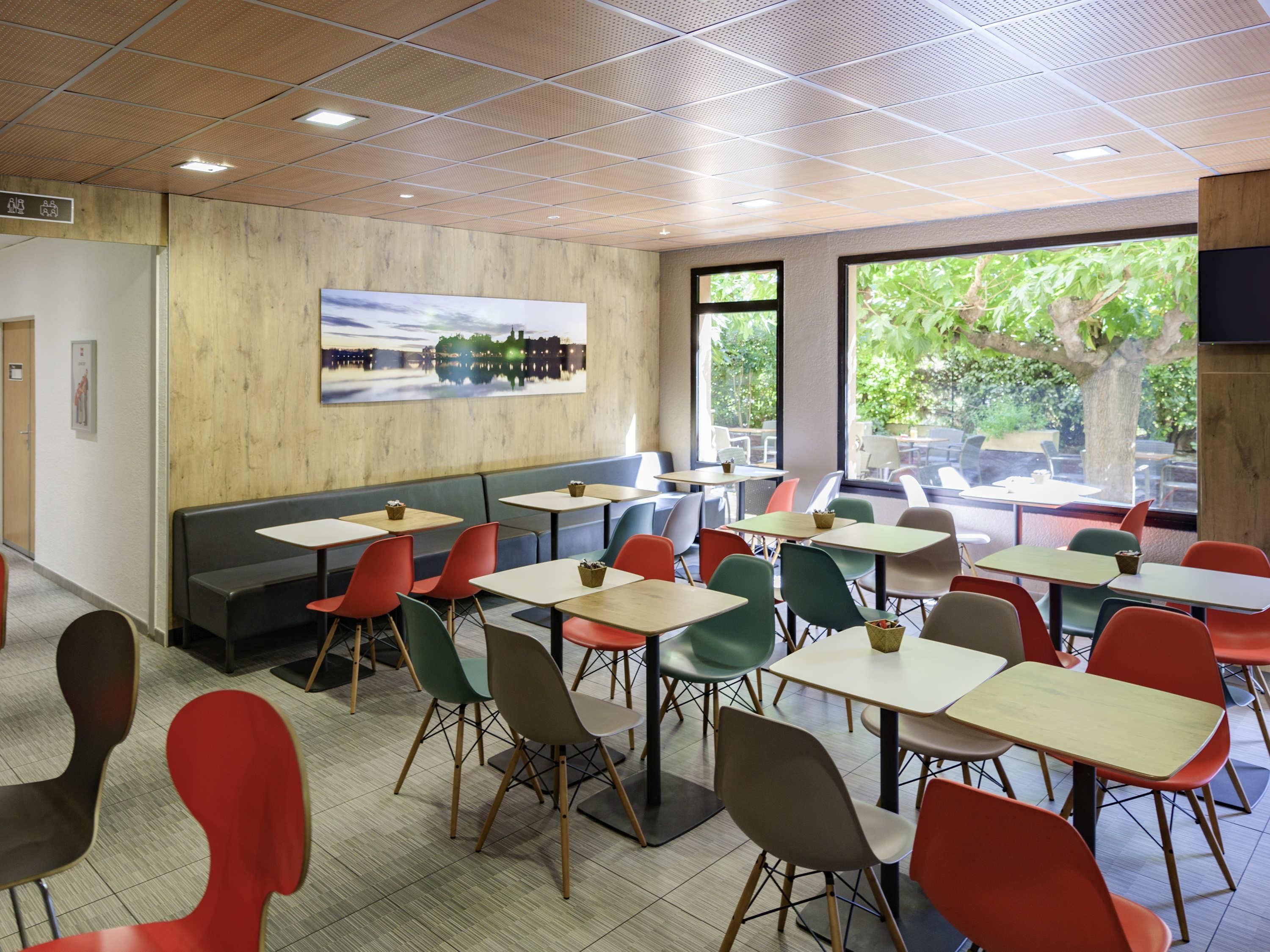 Un espace de restauration moderne, avec des banquettes et des tables, de grandes fenêtres offrant une vue sur la verdure, et une peinture représentant un paysage urbain accrochée au mur.