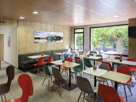 Un comedor moderno con cabinas y mesas, amplias ventanas con vistas a zonas verdes, y un cuadro de paisaje urbano en la pared.