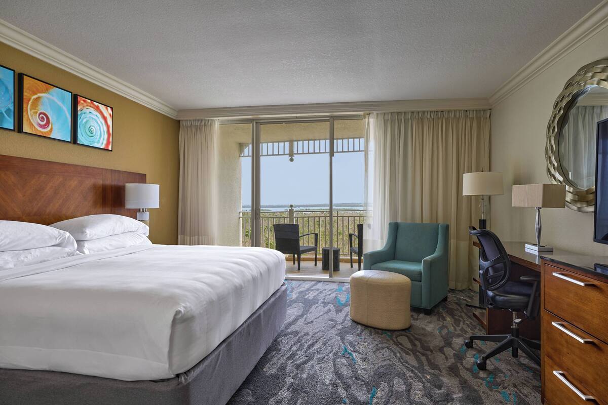 A hotel room with a large bed, a desk with a chair, a small ottoman, and a balcony with a view.