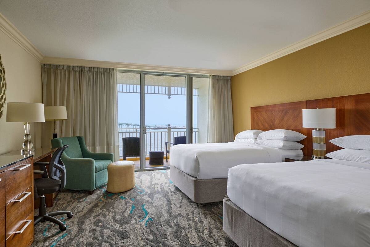 A hotel room with two beds, a desk, a chair, a sofa, and a balcony with a view.