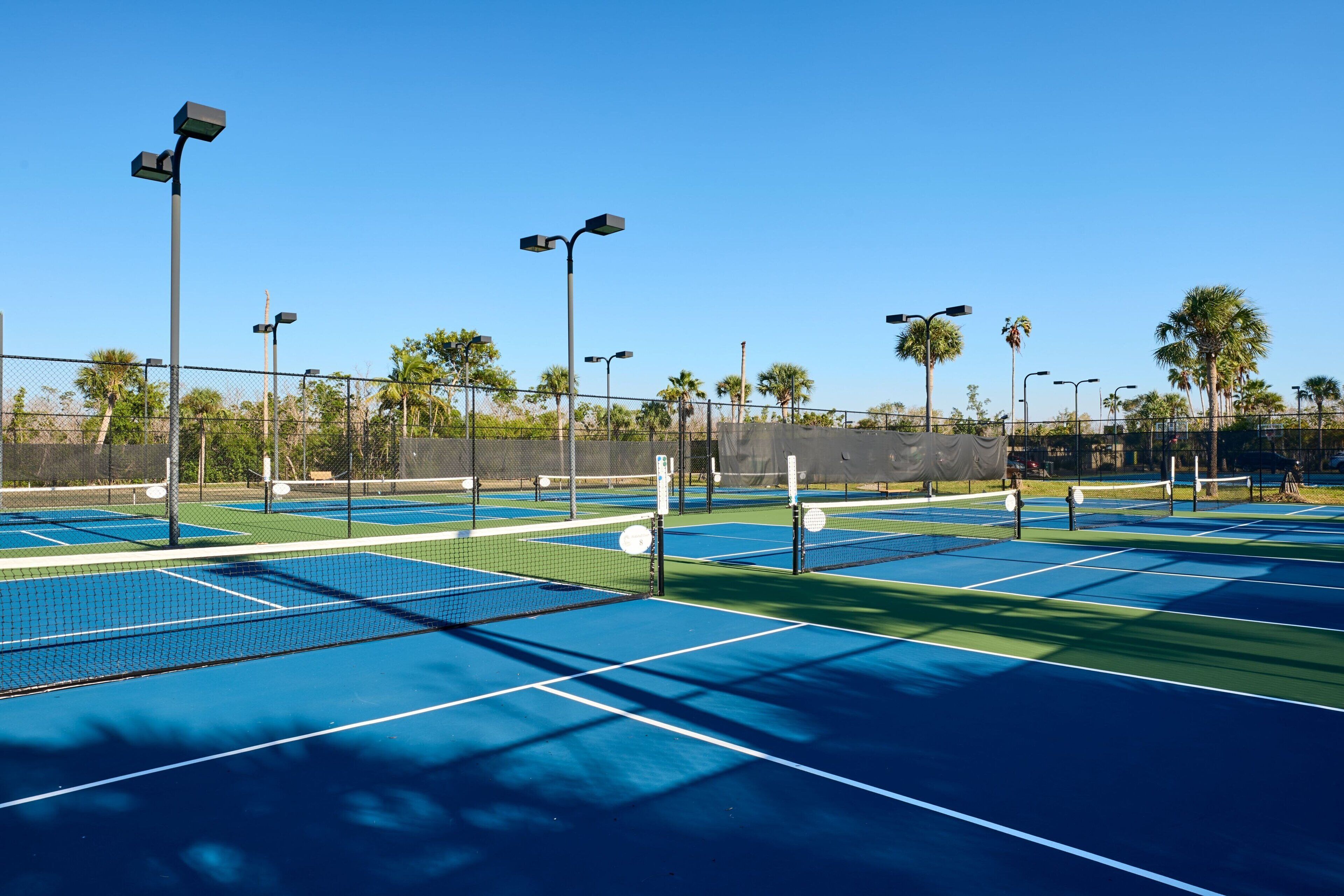 A tennis court with multiple courts, surrounded by a fence, and equipped with street lamps.