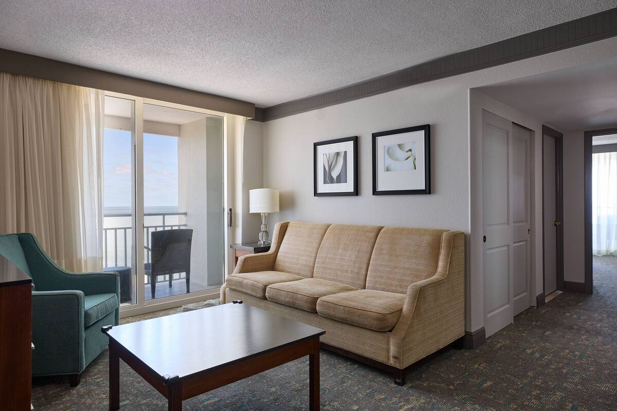 A hotel room with a sofa, a chair, a table, a lamp, and two framed pictures on the wall.