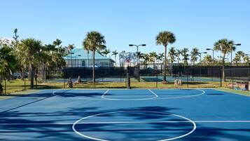 An outdoor basketball court with clear markings, surrounded by palm trees and a fence.